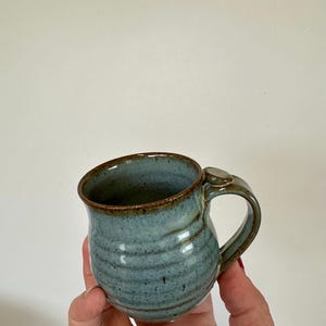 May include: A handmade ceramic mug with a blue-green glaze and a brown rim. The mug has a curved handle and a textured, ridged surface. The mug is held in a hand against a plain white background.