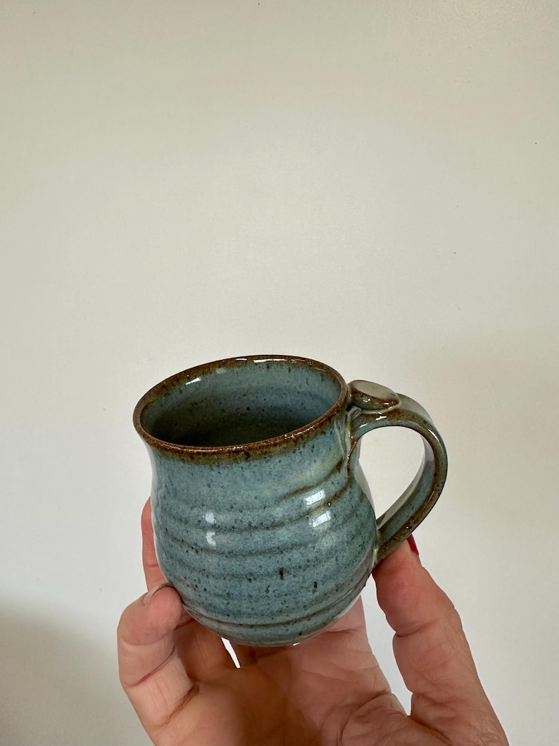 May include: A handmade ceramic mug with a blue-green glaze and a brown rim. The mug has a curved handle and a textured, ridged surface. The mug is held in a hand against a plain white background.