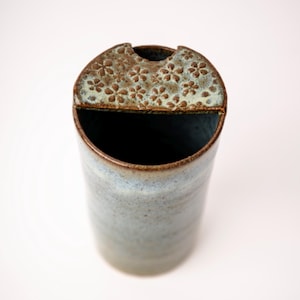 May include: A cylindrical ceramic vessel with a mottled blue and brown glaze. The top features a semi-circular lid with a textured surface decorated with a repeating pattern of small, impressed flower shapes in a light blue and brown finish.