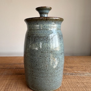 May include: A ceramic jar with a lid, featuring a speckled blue glaze with brown accents. The jar has a rounded body and a slightly flared opening.