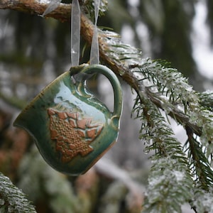 May include: A handmade, green ceramic mug ornament with a brown embossed design, hanging from a tree branch. The mug has a handle and is suspended by a clear ribbon. The background shows icy evergreen needles.