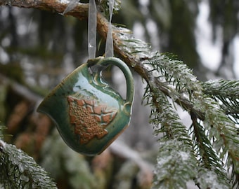 May include: A handmade, green ceramic mug ornament with a brown embossed design, hanging from a tree branch. The mug has a handle and is suspended by a clear ribbon. The background shows icy evergreen needles.