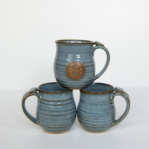 Puede incluir: Tres tazas de cerámica azules apiladas. Cada taza tiene un asa y un borde marrón. La taza superior tiene un emblema marrón. Las tazas son hechas a mano y tienen un aspecto rústico. Altura aproximada de 10 cm.