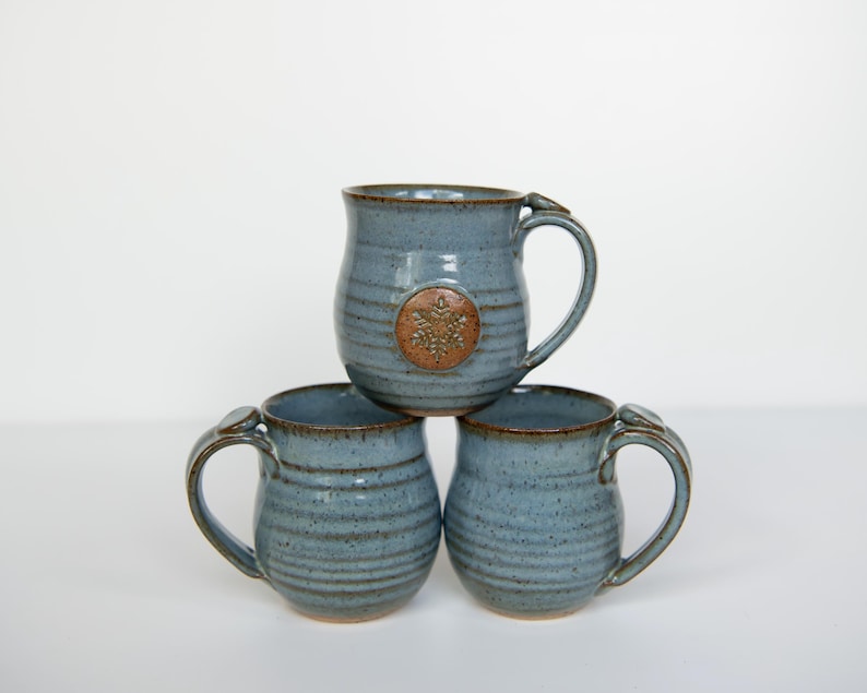 May include: Three blue ceramic mugs stacked on top of each other. Each mug has a handle and a brown rim. The top mug has a brown emblem. The mugs are handmade and have a rustic appearance.