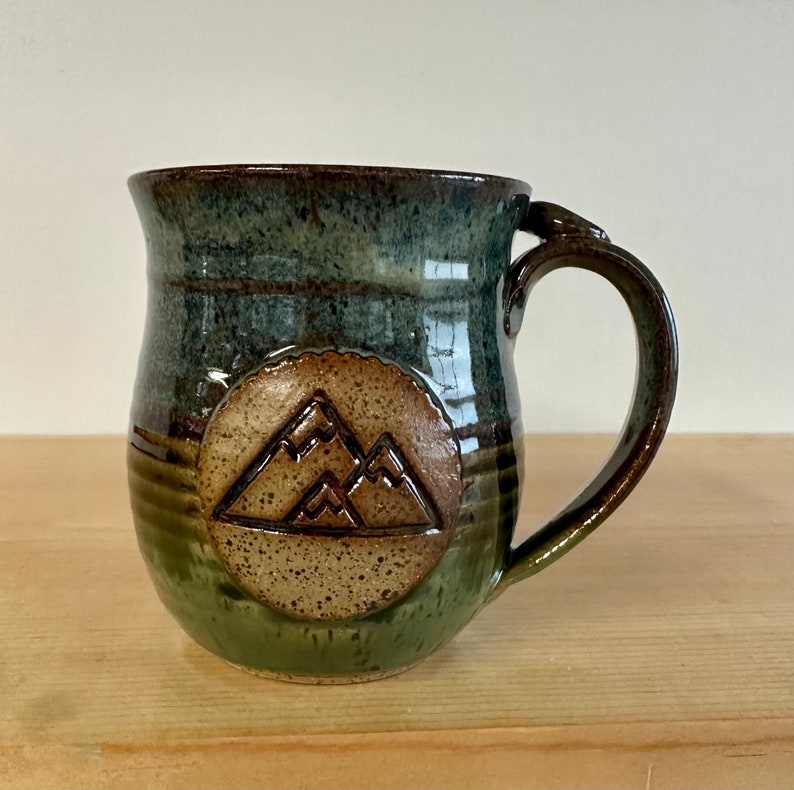 May include: A handmade ceramic mug with a mountain design. The mug features a dark green and brown glaze with a circular mountain emblem. The handle is a dark brown color. The mug is sitting on a wooden surface.