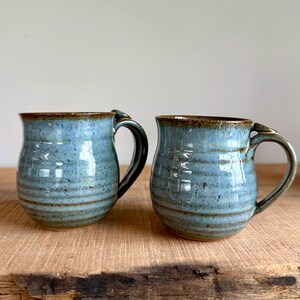 May include: Two handmade ceramic mugs with a blue glaze and brown rims and handles. The mugs have a rounded shape and a speckled texture, perfect for coffee or tea. The mugs are sitting on a wooden surface.