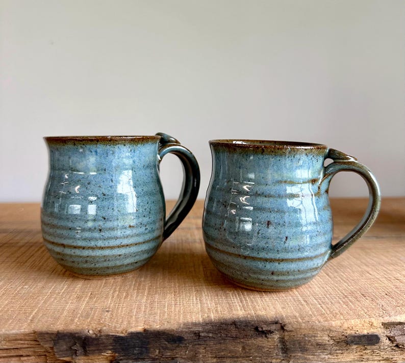 May include: Two handmade ceramic mugs with a blue glaze and brown rims and handles. The mugs have a rounded shape and a speckled texture, perfect for coffee or tea. The mugs are sitting on a wooden surface.