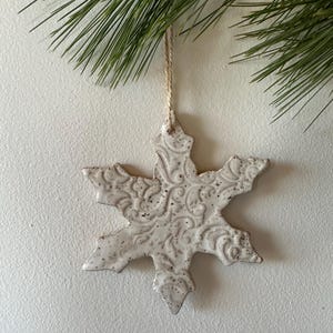 May include: A white ceramic snowflake ornament with a detailed, embossed design. The ornament has a rustic, farmhouse style and is perfect for adding a touch of winter charm to your home decor.