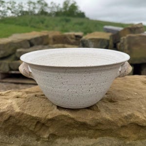 May include: A speckled off-white ceramic bowl with a wide, flared rim and two small, textured handles on opposite sides. The bowl features a subtle, light-colored speckled pattern throughout its surface and a smooth, rounded base.