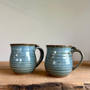 May include: Two handmade ceramic mugs with a blue glaze and brown trim. The mugs have a rounded shape with a handle and are sitting on a wooden surface. The mugs are approximately 4 inches tall.