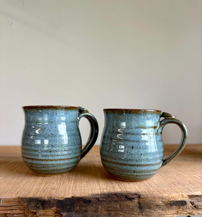 May include: Two handmade ceramic mugs with a blue glaze and brown trim. The mugs have a rounded shape with a handle and are sitting on a wooden surface. The mugs are approximately 4 inches tall.