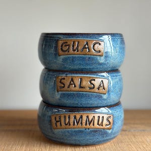 May include: Three blue ceramic bowls with brown glaze stacked on top of each other. The bowls are labeled "GUAC", "SALSA", and "HUMMUS" in black lettering on a brown rectangle.