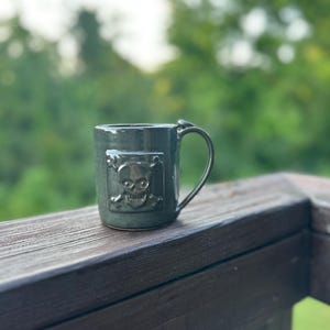 May include: A dark green ceramic mug featuring a raised, square-shaped relief of a skull and crossbones on the front. The mug has a smooth, glossy finish and a curved handle on the right side, providing a sturdy grip for holding hot or cold beverages.