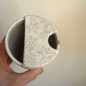 May include: White ceramic mug with a unique design featuring a cutout for a tea bag. The mug has a speckled glaze and a subtle floral pattern.