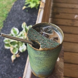 May include: A green ceramic tumbler with a textured, dark green semi-circular lid featuring a small opening for a metal straw. The tumbler contains a light brown iced beverage with visible ice cubes. The exterior has a mottled, earthy finish.