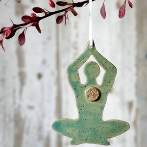 May include: A ceramic ornament in the shape of a person in a yoga pose, with arms raised. The ornament is a speckled teal color with a raised lotus flower design on the chest. It hangs from a white ribbon.