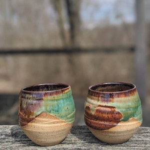 May include: Two ceramic mugs with a brown, green, and beige glaze. The mugs are sitting on a wooden surface.