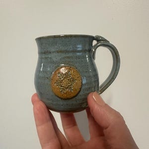 May include: A handmade ceramic mug with a blue-grey glaze. The mug features a circular, raised, tan-colored snowflake design. The handle is a curved loop, and the mug is held in a hand.