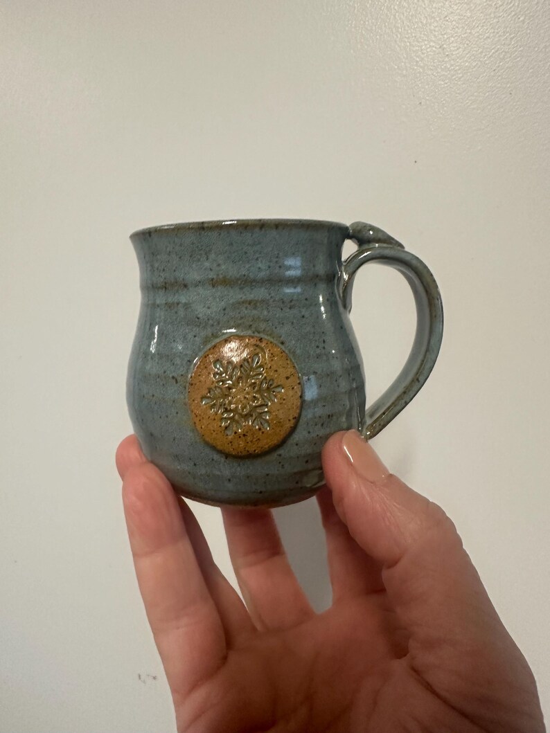 May include: A handmade ceramic mug with a blue-grey glaze. The mug features a circular, raised, tan-colored snowflake design. The handle is a curved loop, and the mug is held in a hand.