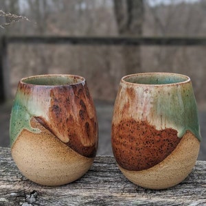 May include: Two ceramic mugs with a brown, green, and beige glaze. The mugs have a rounded shape and a wide base.