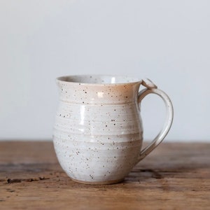 May include: A speckled white ceramic mug with a rounded body and a curved handle. The mug has a light brown rim and is set on a wooden surface. The handmade mug is suitable for coffee, tea, or other beverages.