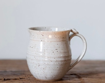 May include: A handmade ceramic mug in speckled white with a rounded shape and a curved handle. The mug has a light brown rim and sits on a wooden surface. Suitable for hot drinks, the mug is a rustic addition to any kitchen.
