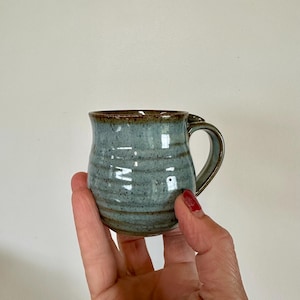 May include: A small, handmade ceramic mug with a blue-green glaze and a brown rim. The mug has a rounded shape with a handle and is held in a person's hand. The mug is approximately 3 inches tall.