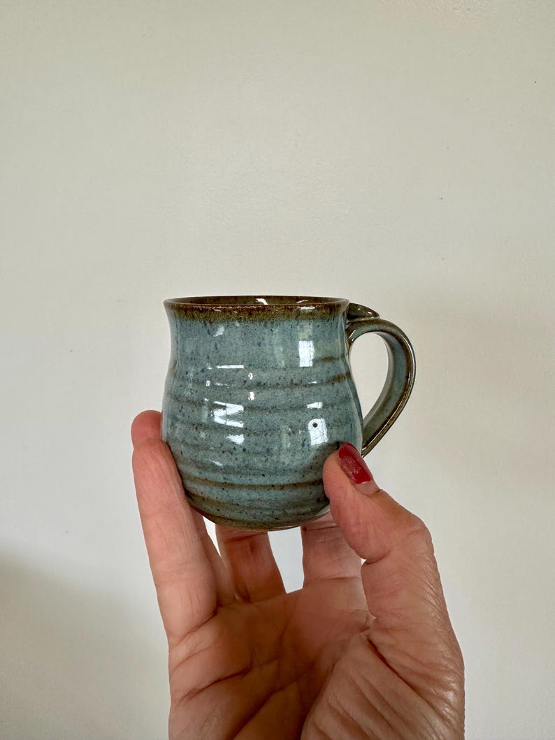 May include: A small, handmade ceramic mug with a blue-green glaze and a brown rim. The mug has a rounded shape with a handle and is held in a person's hand. The mug is approximately 3 inches tall.