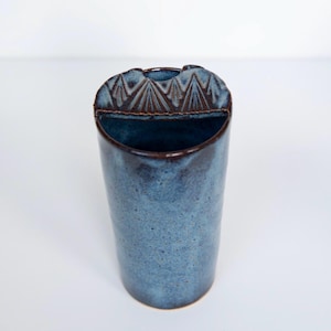 May include: A cylindrical ceramic vase with a mottled blue glaze and a dark brown rim. The top edge features a semi-circular extension with a carved geometric pattern of repeating triangles. The interior is dark, and the base is rounded.