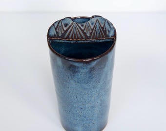 May include: A cylindrical ceramic vase with a mottled blue glaze and a dark brown rim. The top edge features a semi-circular extension with a carved geometric pattern of repeating triangles. The interior is dark, and the base is rounded.