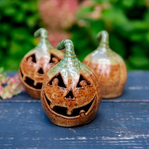 May include: Three ceramic pumpkin-shaped lanterns with a glossy finish. Each piece features a speckled brown body, a green stem, and carved triangular eyes and a wide, toothy grin. The hollow structures allow light to pass through the cutouts.