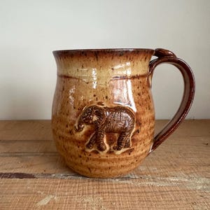 May include: A handmade ceramic mug with a brown elephant relief. The mug has a rounded shape with a large handle and a speckled, earthy brown glaze. The elephant is detailed and centered on the mug's body.