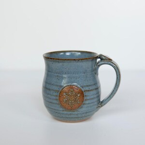 May include: A handmade ceramic mug in a speckled blue glaze. The mug features a brown snowflake design and a curved handle. The mug is a unique piece of pottery, perfect for enjoying coffee or tea.