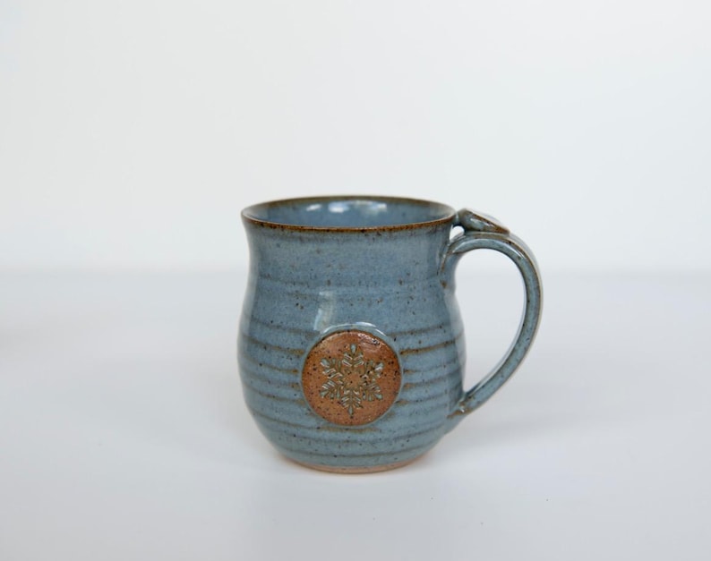 May include: A handmade ceramic mug in a speckled blue glaze. The mug features a brown snowflake design and a curved handle. The mug is a unique piece of pottery, perfect for enjoying coffee or tea.