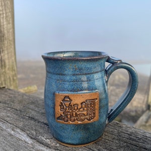 May include: A blue ceramic mug with a brown train design. The mug has a large handle and a textured surface. The train design is embossed on a rectangular patch on the mug's side. The mug is sitting on a wooden surface.