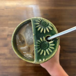 May include: Overhead view of a green ceramic cup with a floral design on the lid, holding iced coffee and a metal straw. The cup is divided into two sections. The background is a wooden surface.