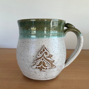 May include: A handmade ceramic mug with a white speckled finish and a green rim. The mug features a raised brown Christmas tree design on the front and a decorative handle. This mug is perfect for enjoying hot beverages.