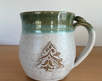 Op de afbeelding: Handgemaakte keramische mok met een witte gespikkelde afwerking en een groene rand. De mok heeft een verhoogd bruin kerstboomontwerp aan de voorkant en een decoratief handvat. Perfect voor warme dranken.