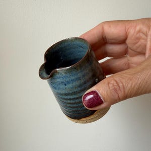 May include: A small ceramic creamer pitcher featuring a deep blue and dark brown reactive glaze with horizontal ridges along the body. The base is made of unglazed, speckled beige clay, and the rim includes a small, functional pouring spout.