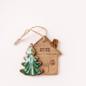 May include: A ceramic Christmas ornament featuring a house and a Christmas tree. The house is tan with the year "2025" and a chimney. The tree is green. A twine loop is attached for hanging.