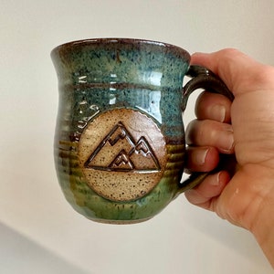 May include: A handmade ceramic mug with a mountain design. The mug features a brown handle and a round emblem with a mountain range. The mug has a blue and green glaze, with a brown rim and base. The mug is held by a hand.