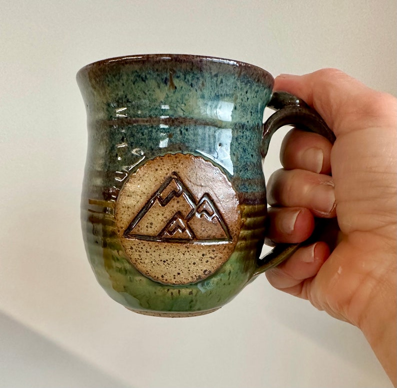May include: A handmade ceramic mug with a mountain design. The mug features a brown handle and a round emblem with a mountain range. The mug has a blue and green glaze, with a brown rim and base. The mug is held by a hand.