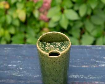 May include: A cylindrical ceramic travel mug in a speckled olive green glaze. The top features a semi-circular lid section with a raised, embossed pattern of small monstera leaves and a central circular opening for drinking or a straw.
