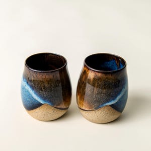 May include: Two stemless ceramic wine glasses with a unique glaze. The glasses have a dark brown interior and a gradient exterior, transitioning from a light tan base to blue and brown hues. The rim is dark brown.