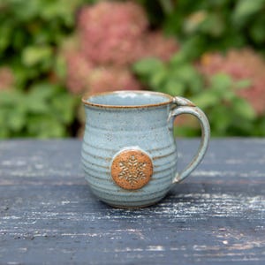 May include: A handmade ceramic mug with a light blue glaze and a brown snowflake design. The mug has a rounded shape with a handle and is approximately 4 inches tall.