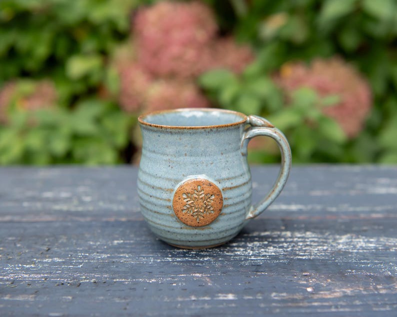May include: A handmade ceramic mug with a light blue glaze and a brown snowflake design. The mug has a rounded shape with a handle and is approximately 4 inches tall.