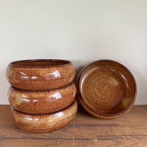 May include: A stack of four handmade ceramic bowls with a speckled, glossy brown glaze. The bowls are round with a wide rim and sit on a wooden surface. One additional bowl is placed to the right.