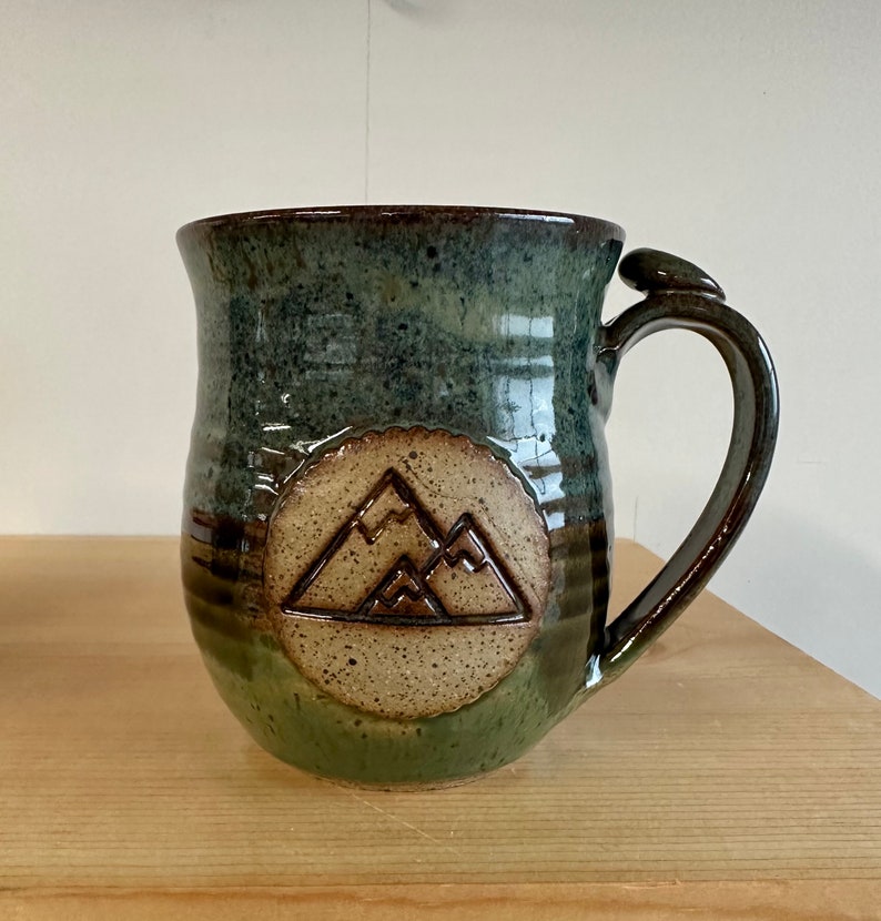 May include: A handmade ceramic mug with a mountain design. The mug features a dark green and brown glaze, with a circular mountain emblem on the front. The handle has a unique, curved design. This mug is perfect for coffee or tea.