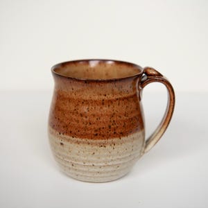 May include: A handmade ceramic mug with a rounded body and a curved handle. The mug features a two-tone design, with a light beige base and a speckled brown upper section. The rim is a darker brown.