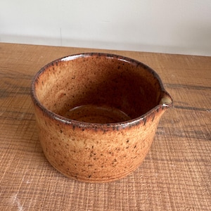 May include: A handmade, speckled ceramic bowl in shades of brown. The bowl has a rounded shape with a small spout and a dark brown rim. The interior is a lighter brown, and the exterior has a textured, speckled appearance.